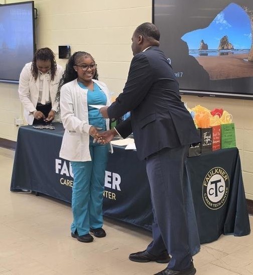 A Faulkner student in scrubs accepts a certificate from Principal Dr. Jason Lafitte