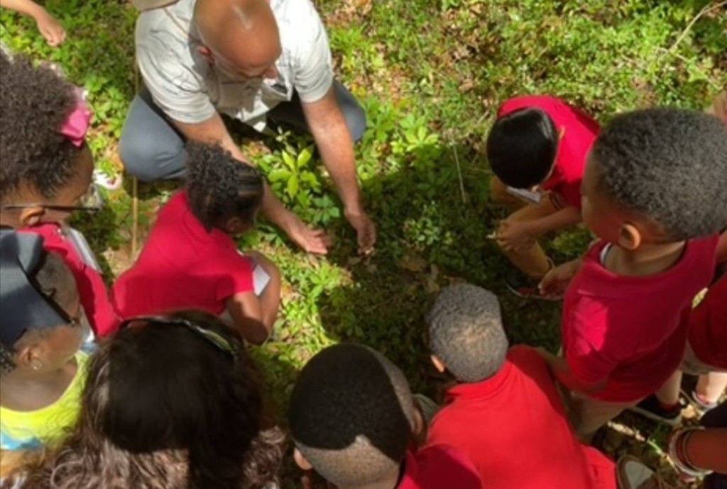 A man shows something in the dirt to a  group of kindergarteners