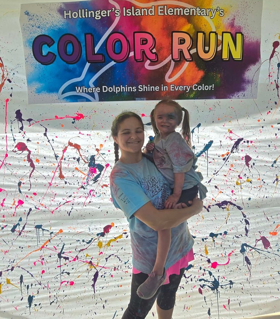 An older girl holds a young girl with colorful chalk on her face, beneath a sign that reads "Color Run"