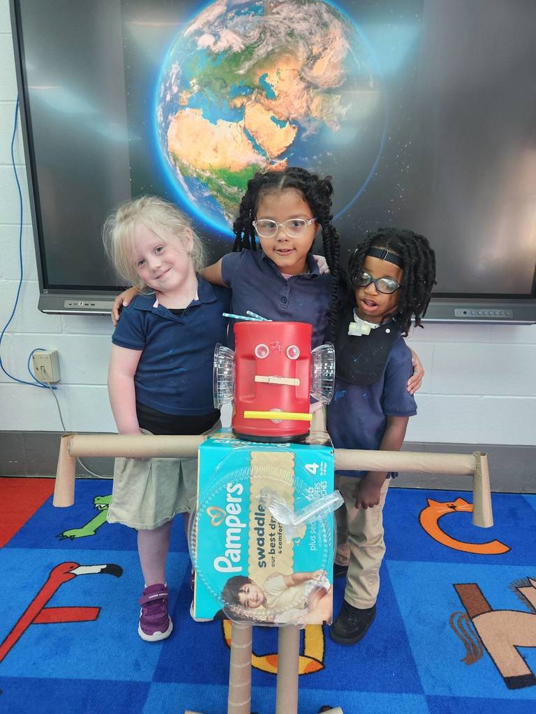 Three Callahan students stand with a robot made from paper towel rolls, a coffee cannister and a diaper box