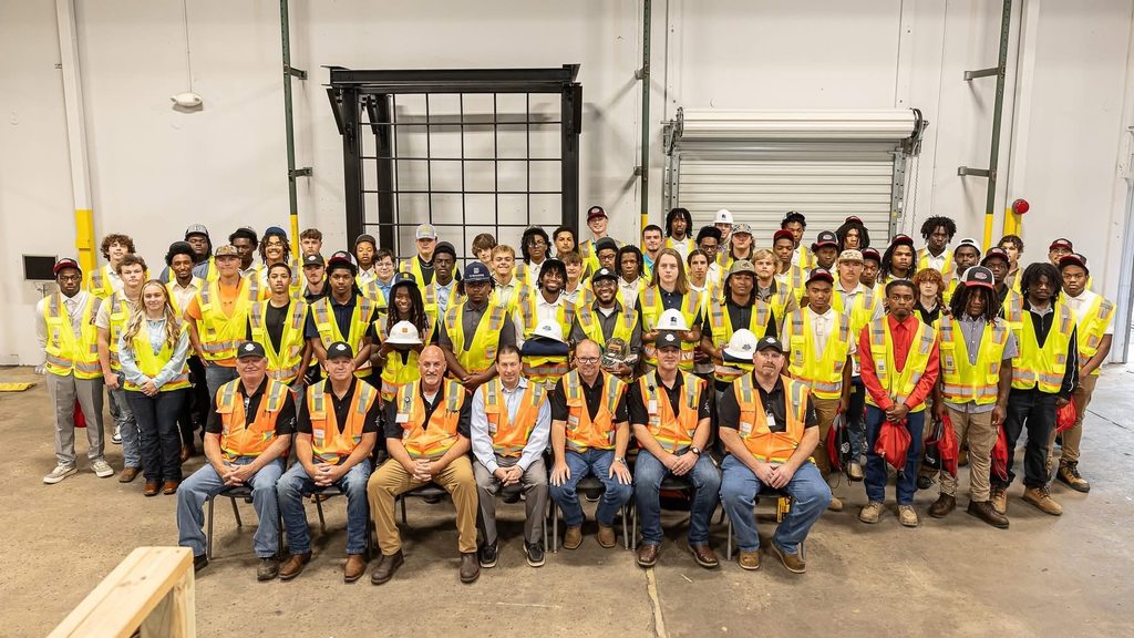 A group of students in reflective vests in a warehouse, with a row of instructors sitting in front of them