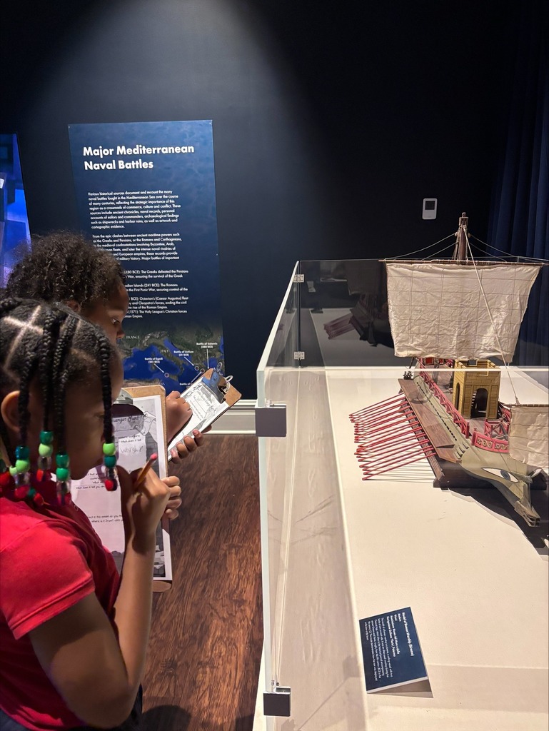 Students from Morningside Elementary standing with clipboards in front of a history display about naval battles  as they take notes.