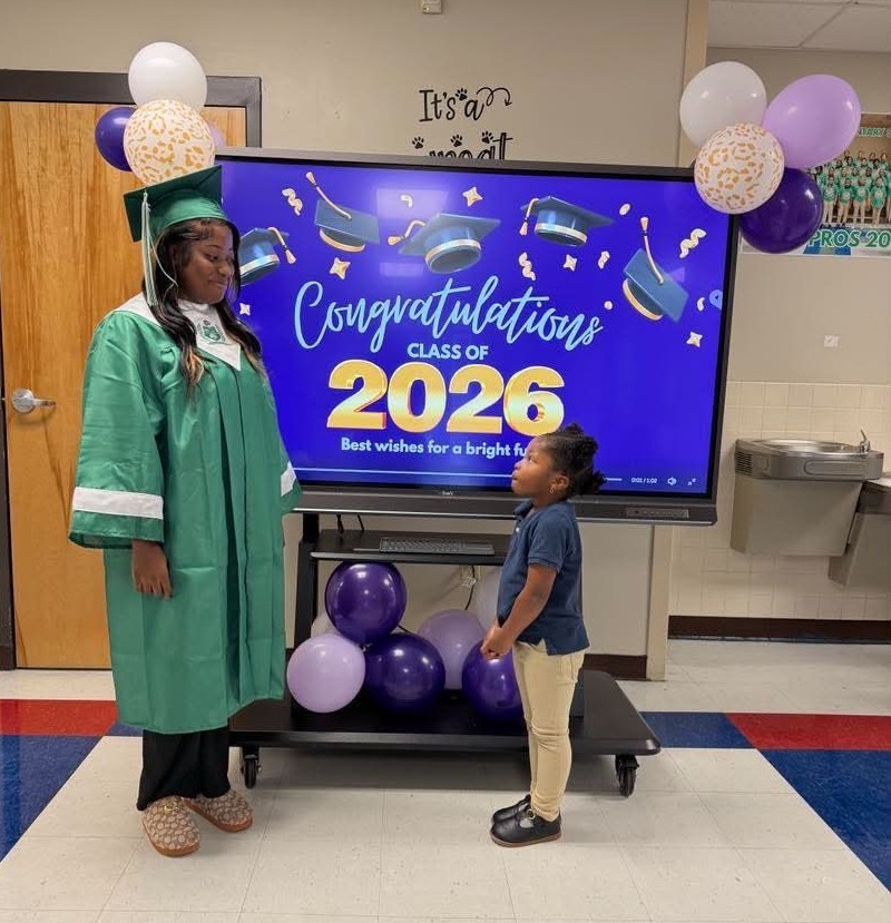 A senior in a green cap and gown stands with an elementary student, who is looking up at her