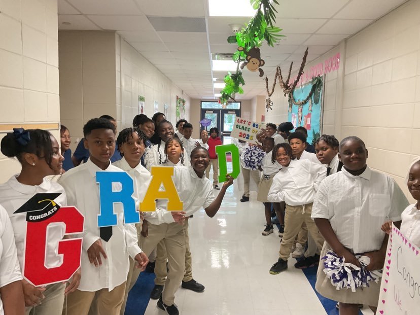 Elementary students stand in a hallway with pompoms and the cutout letters GRAD