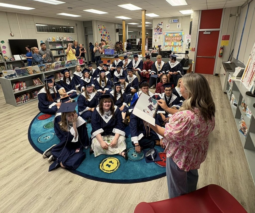 A group of seniors in caps and gowns sitting on the floor of a library as a teacher reads a story