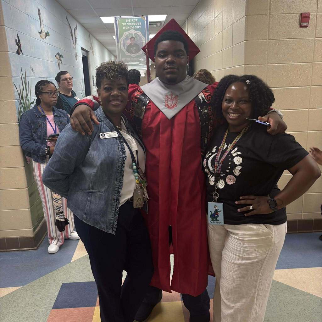 A senior in a red cap and gown hugs a principal and teacher at Gilliard Elementary
