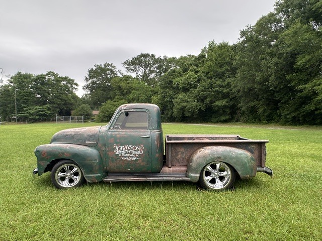 An old, rusted pickup truck with chrome rims parked on a grassy field with green trees in the background.