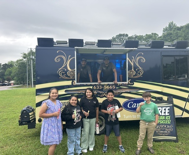 Five children stand in a grassy field, smiling for a photo in front of a black-and-yellow food truck.