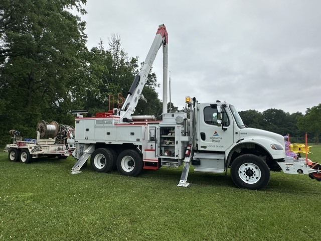 White utility truck with extended crane parked on green grass with trees in the background.