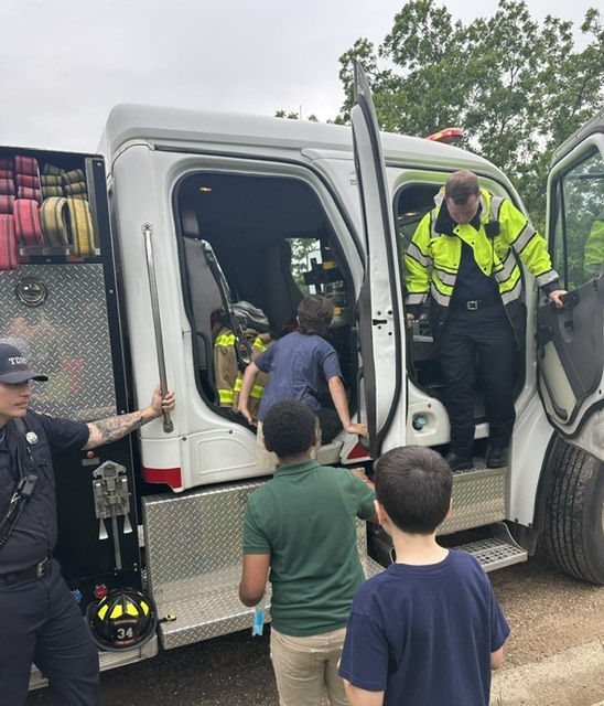 Two firefighters and three children are standing around a fire truck. One child is inside the truck.
