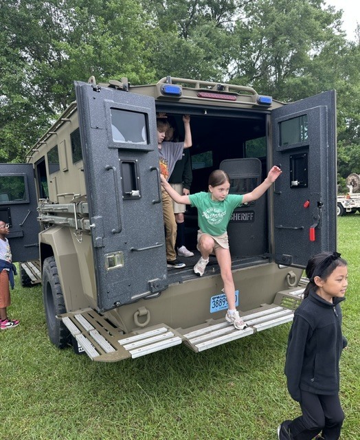 Children exploring an armored vehicle at an outdoor event. One child stands inside the vehicle, while another walks nearby.