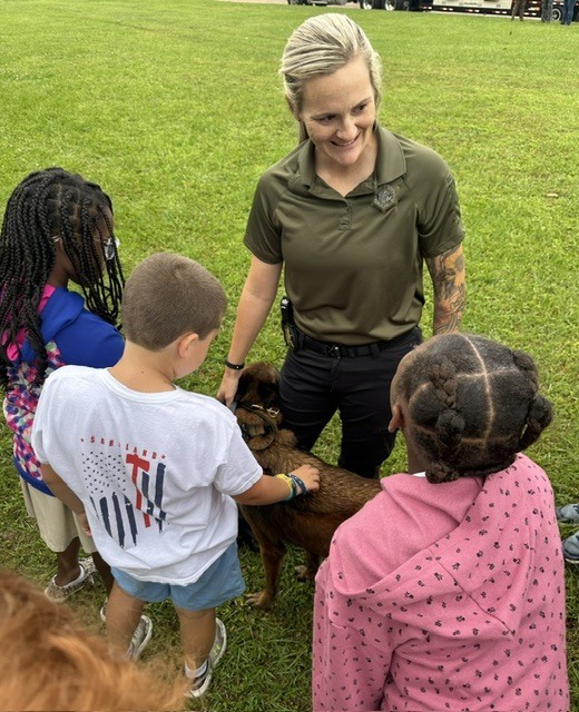 A woman with tattoos, wearing a green shirt, stands with children near a dog on green grass.