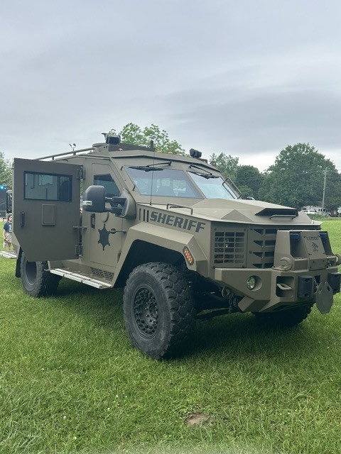 An armored military vehicle, with doors open, is parked on green grass with trees in the background.