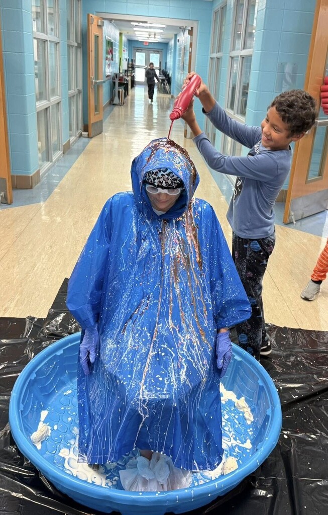 A boy pours chocolate syrup onto the head of Principal Jennifer Fetner, who is wearing a poncho and goggles and sitting in a kiddie pool