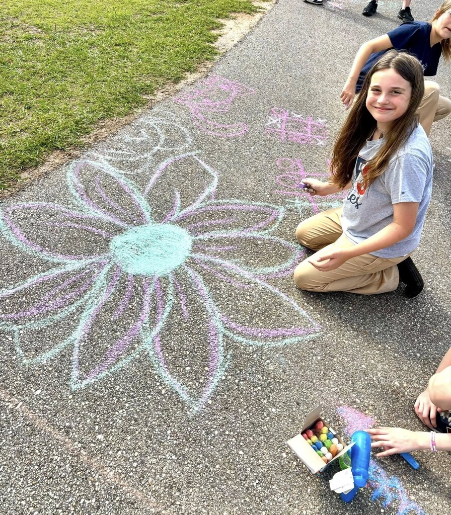 A girl sits by a flower she has drawn on the sidewalk in chalk