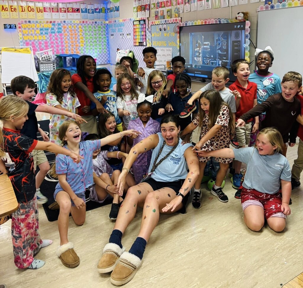 A class of smiling children points to a teacher who is covered in temporary tattoos