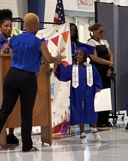 A Maryvale kindergarten student in a blue cap and gown accepts a certificate from a teacher