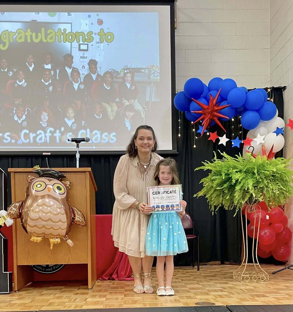 A kindergartener holds a certificate while standing on stage with an Old Shell Road teacher