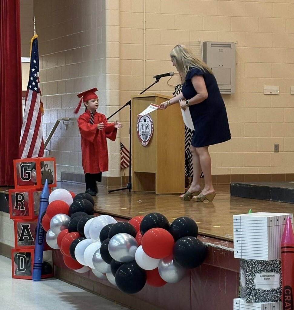 A kindergarten student in a red cap and gown walks on a stage to get a certificate being handed out by a principal