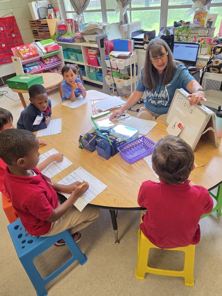 A teacher points to letters on a whiteboard while students around a table watch