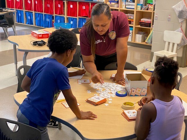 A teacher works with students at a table, using flashcards and letters