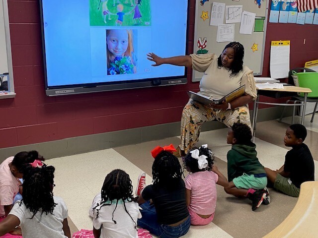 A teacher holds a book and points to a smartboard, while students on the floor watch