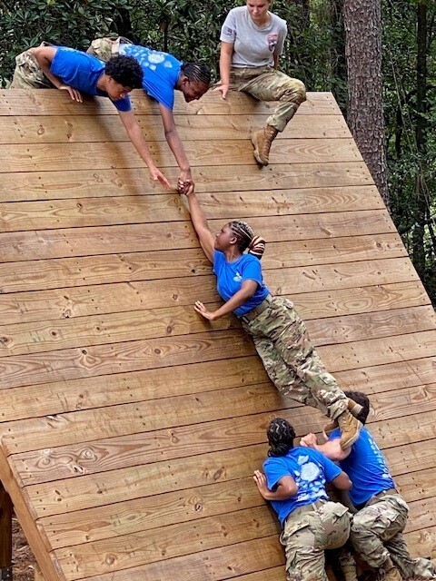 Two JROTC cadets work as a base to help a third climb a wall, while two other cadets reach out their hands at the top