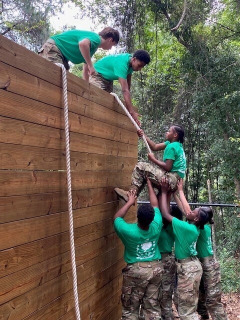 JROTC cadets try to help a fellow cadet climb a rope up a wall