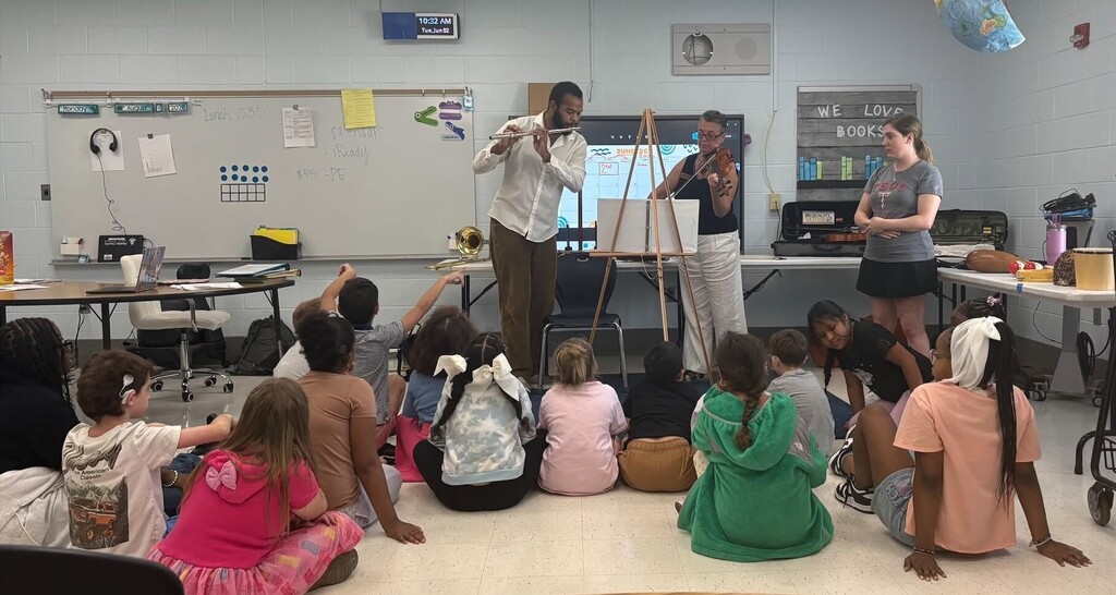A man plays a flute and a woman plays a violin in front of a classroom of children