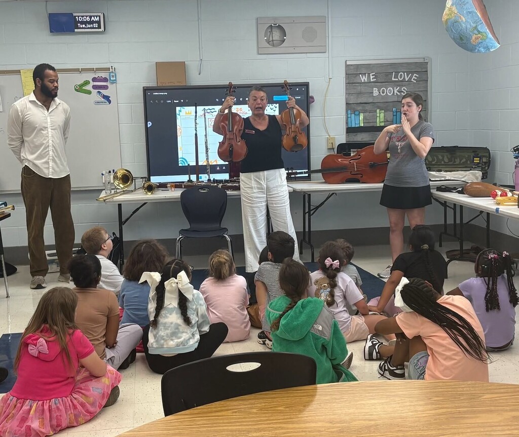 A woman holds up a violin and a viola in front of a classroom of children