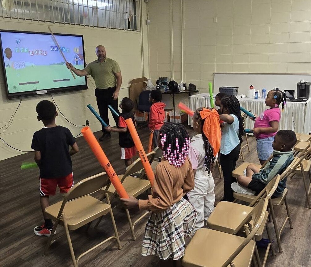 A teacher points to letters on a smartboard, as students hold foam noodles