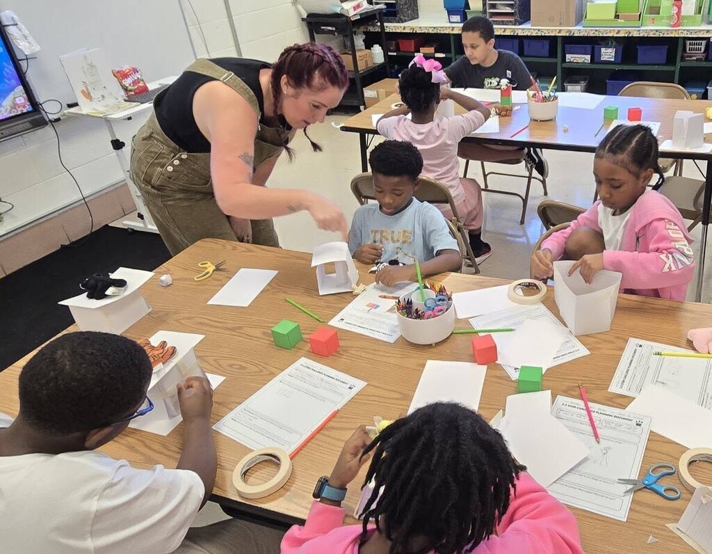 A teacher helps a student as other students around table build towers out of paper