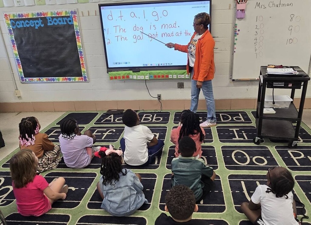 A teacher stands in front of a class of kids, pointing to the sentence "The dog is mad" on a smartboard