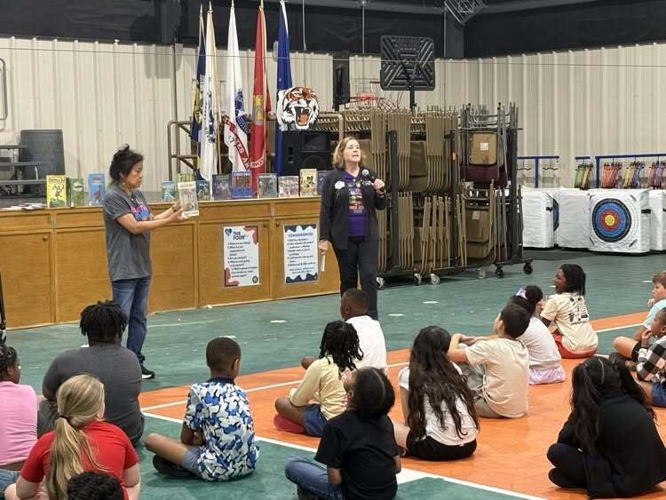 Two women showing books to a group of students sitting on the floor in a gymnasium