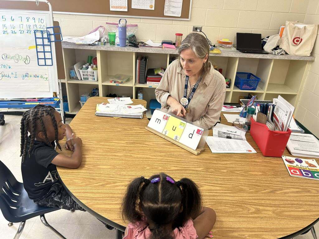 A teacher at a table works on the phonics of the word "mud" with two students