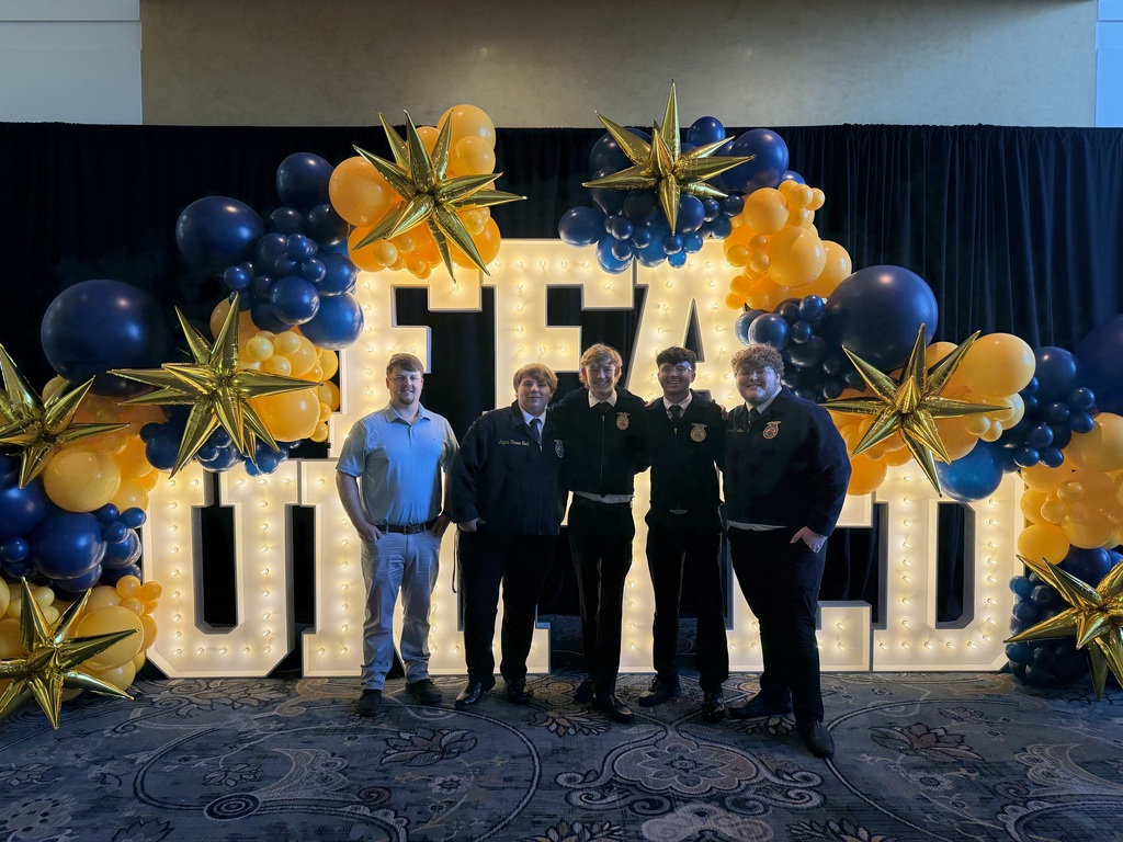A teacher and four high school boys stand in front of a lit FFA sign