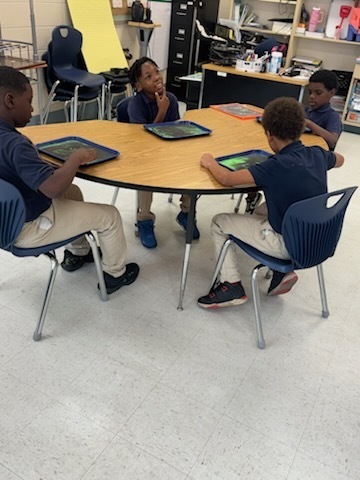 Four children at a table, two holding tablets. Behind them, shelves with books and a yellow board.