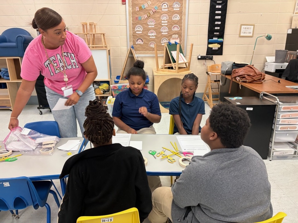 A teacher in a pink shirt leads a discussion with students in a classroom, tables and chairs in the background.