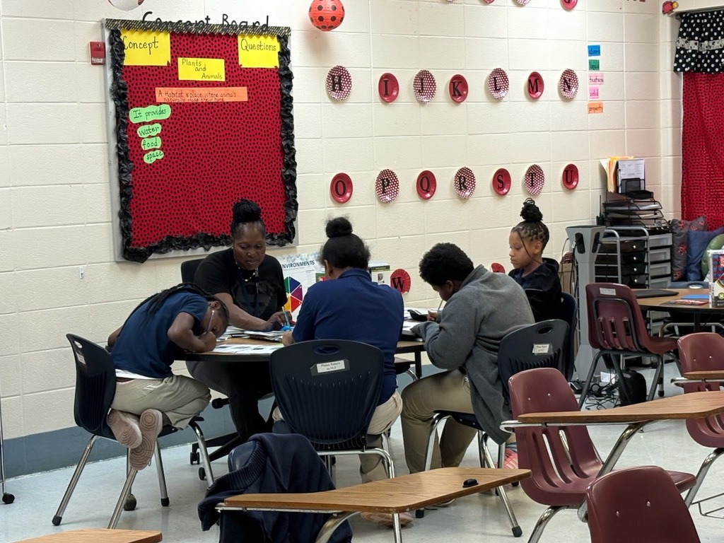 A classroom with students seated at tables. A bulletin board with a red background is on the wall.