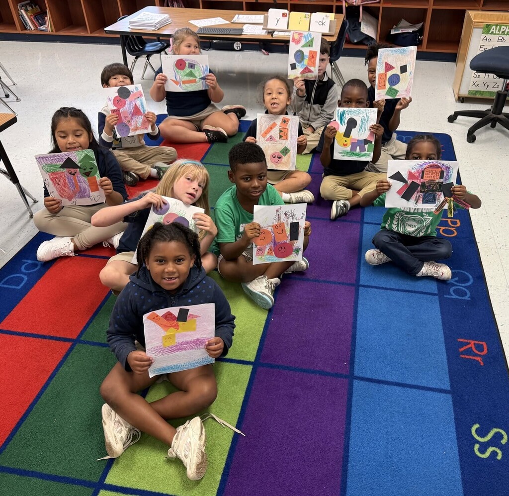 Elementary students sitting on a classroom rug, holding up their artwork