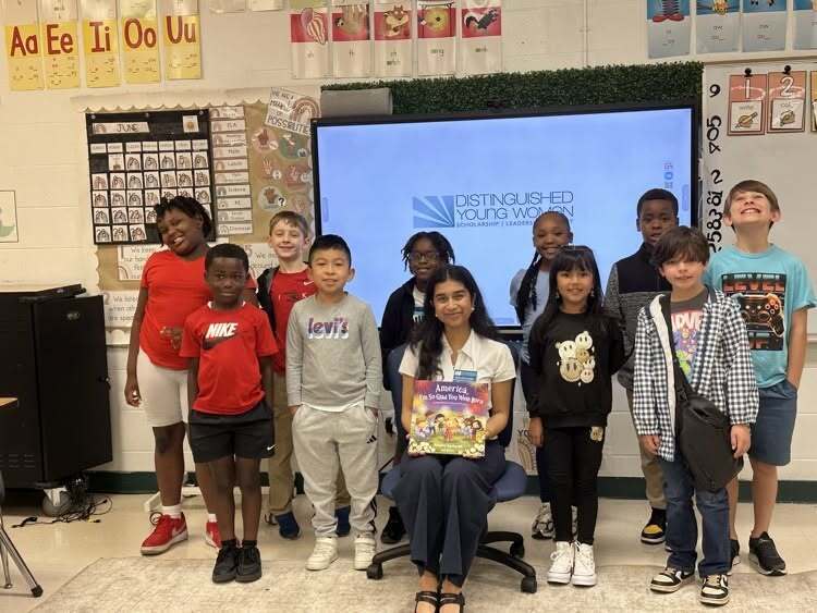 Nikita Chincholkar sits with a book, while several elementary students stand alongside her