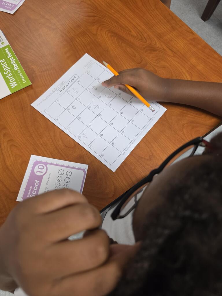 A person wearing glasses sits at a wooden table, working on a worksheet with a pencil.