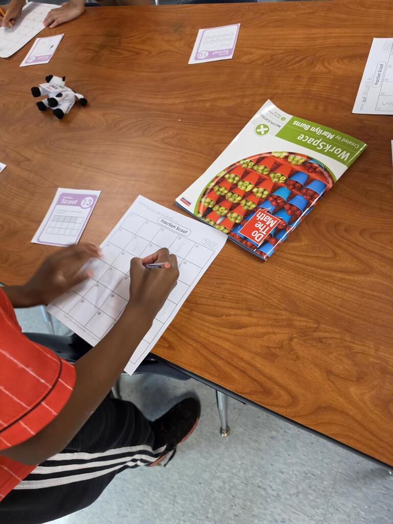 A student sits at a table writing on paper with a pen, surrounded by a workbook and papers.