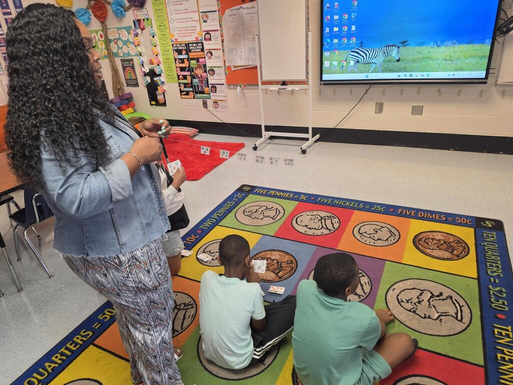 Woman with curly hair stands by large screen. Two boys sit on mat. One boy wears green shirt.