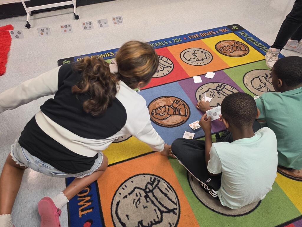 A teacher and two children play a game on a colorful rug with coins. The children sit while the teacher kneels.