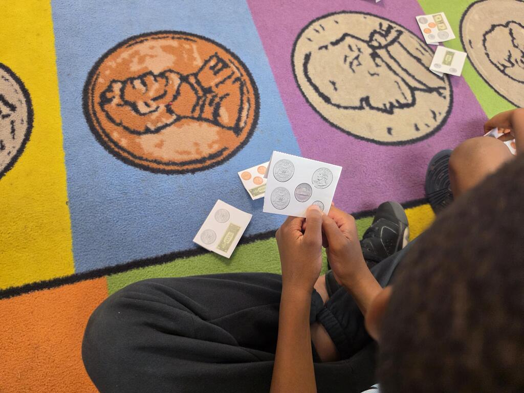 Person seated on floor using cards and coins to teach or learn money. Bright rug has coins and faces.
