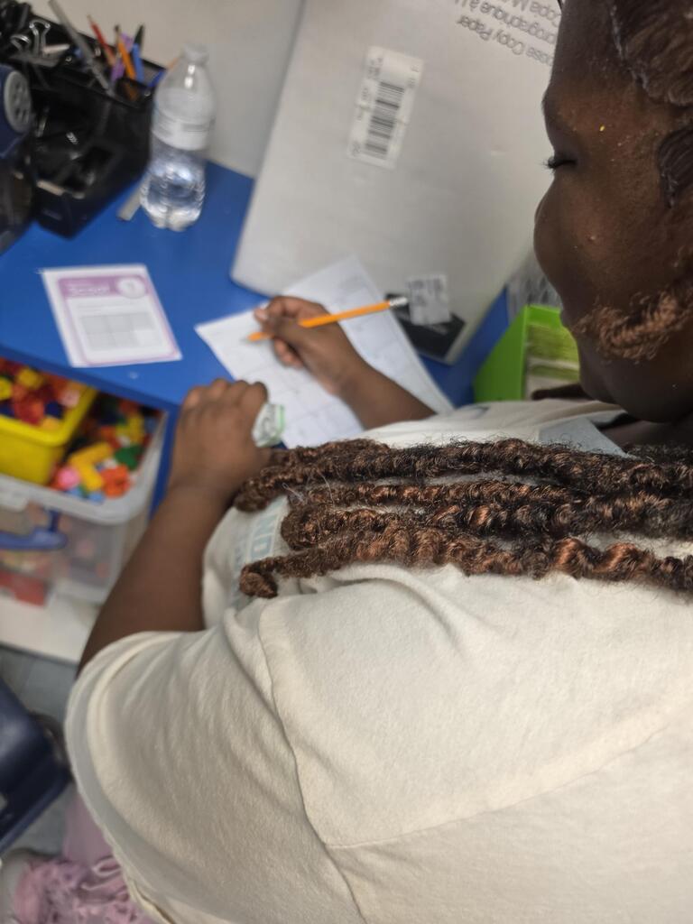Person with braided hair writing on a paper with a pencil at a blue desk.