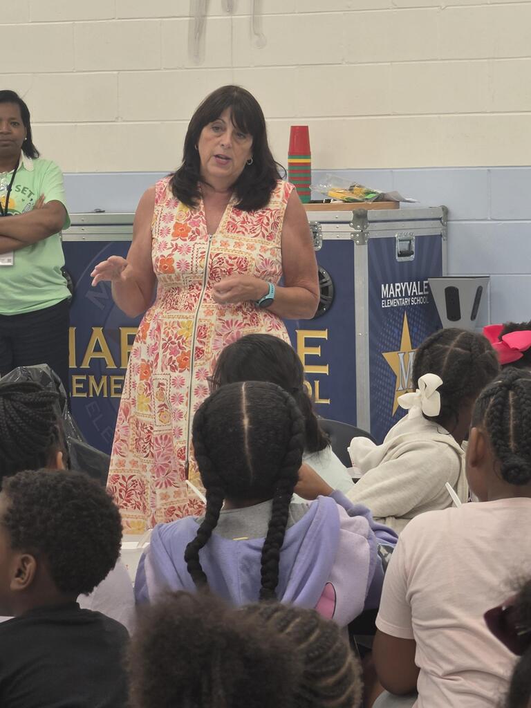 A woman in a floral dress stands before a group of children seated on the floor in a classroom.