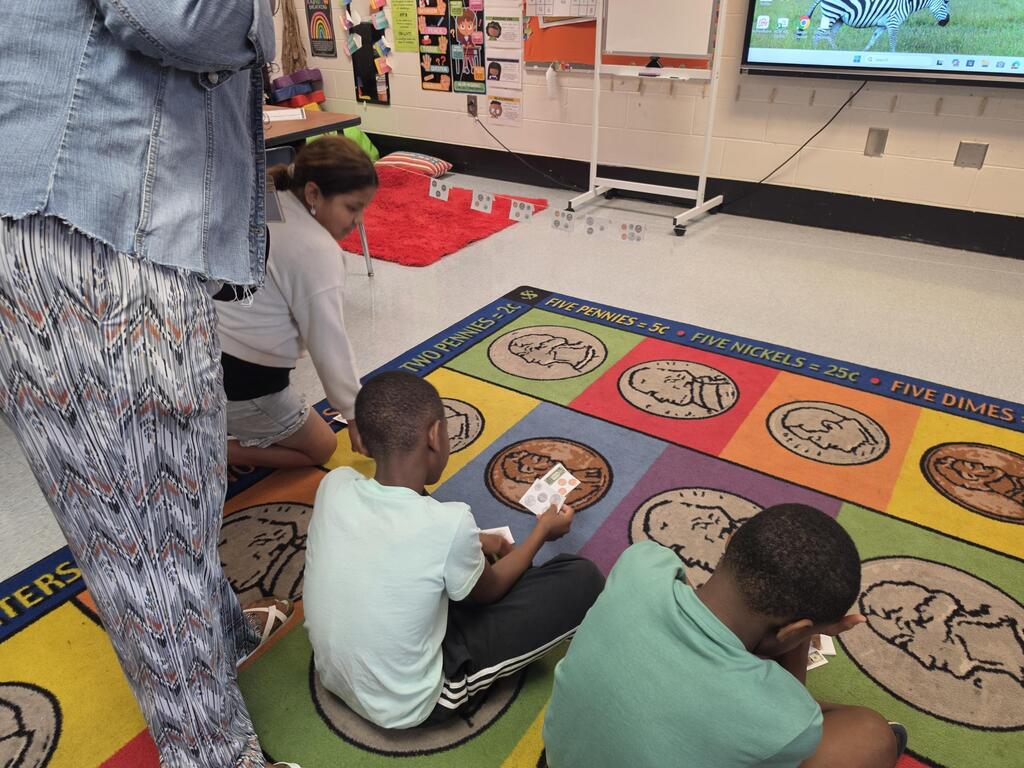 Three children seated on a colorful floor mat playing a game with cards. A woman stands behind them. A monitor on the wall displays a scene.