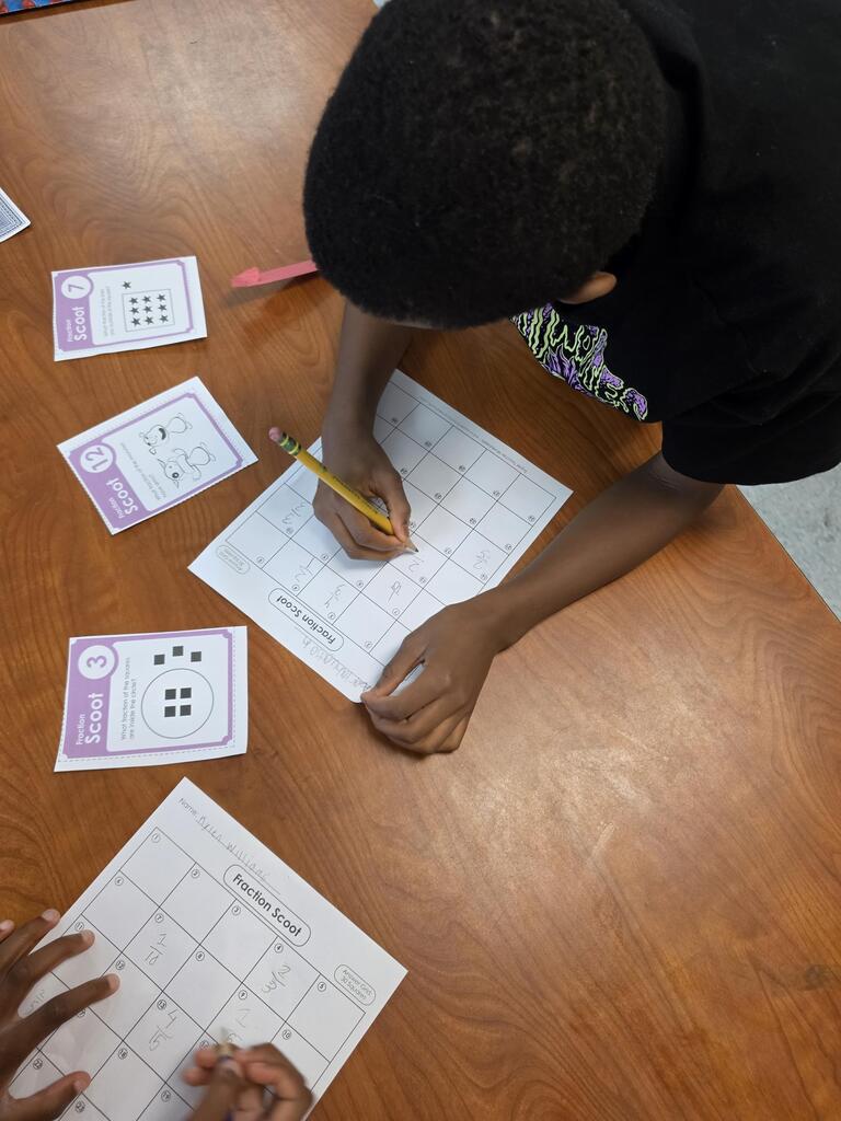 A child completes a worksheet at a table, with several sheets and a pencil in hand.